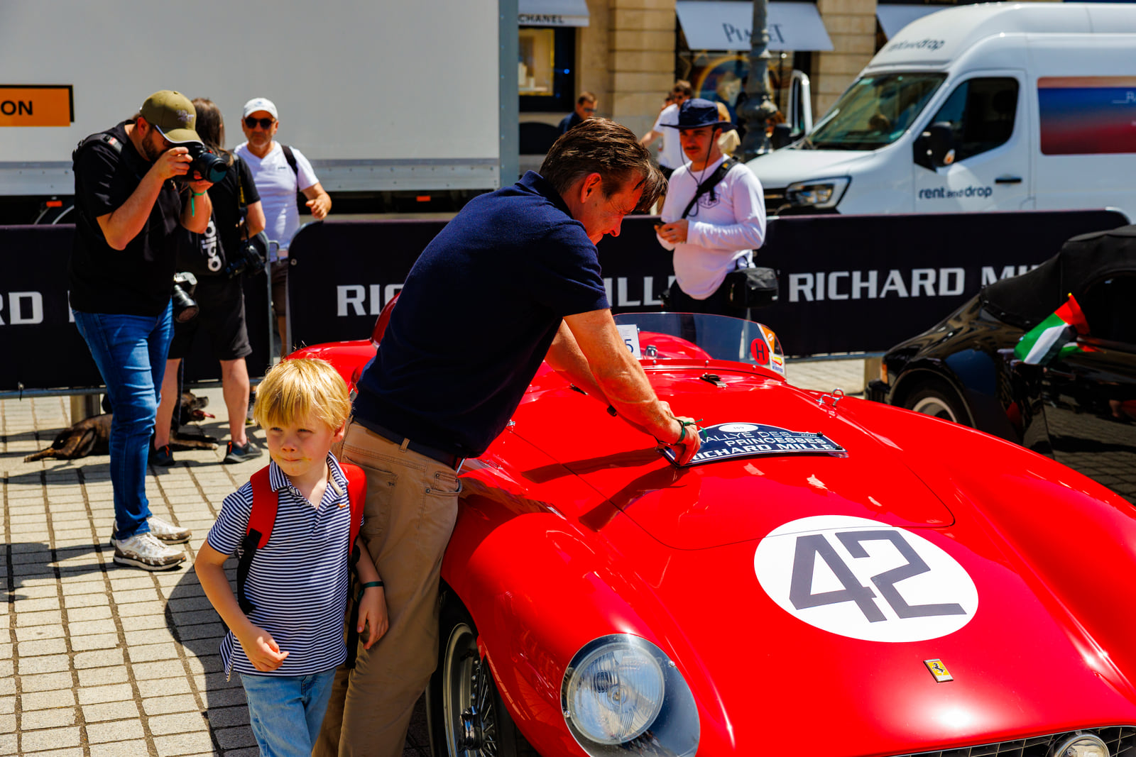 A child beside a vivid red vintage race car on a city street.