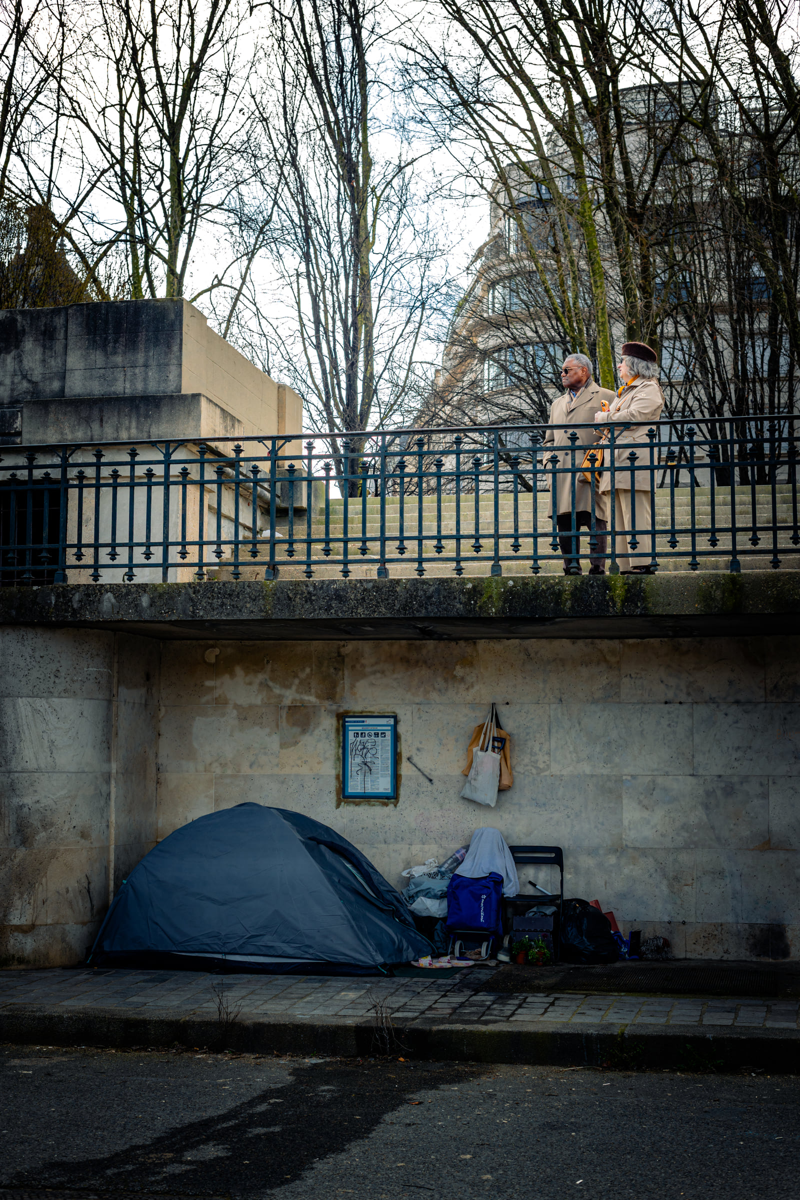 A blue tarpaulin shelter tucked beneath bare trees beside a pale building.