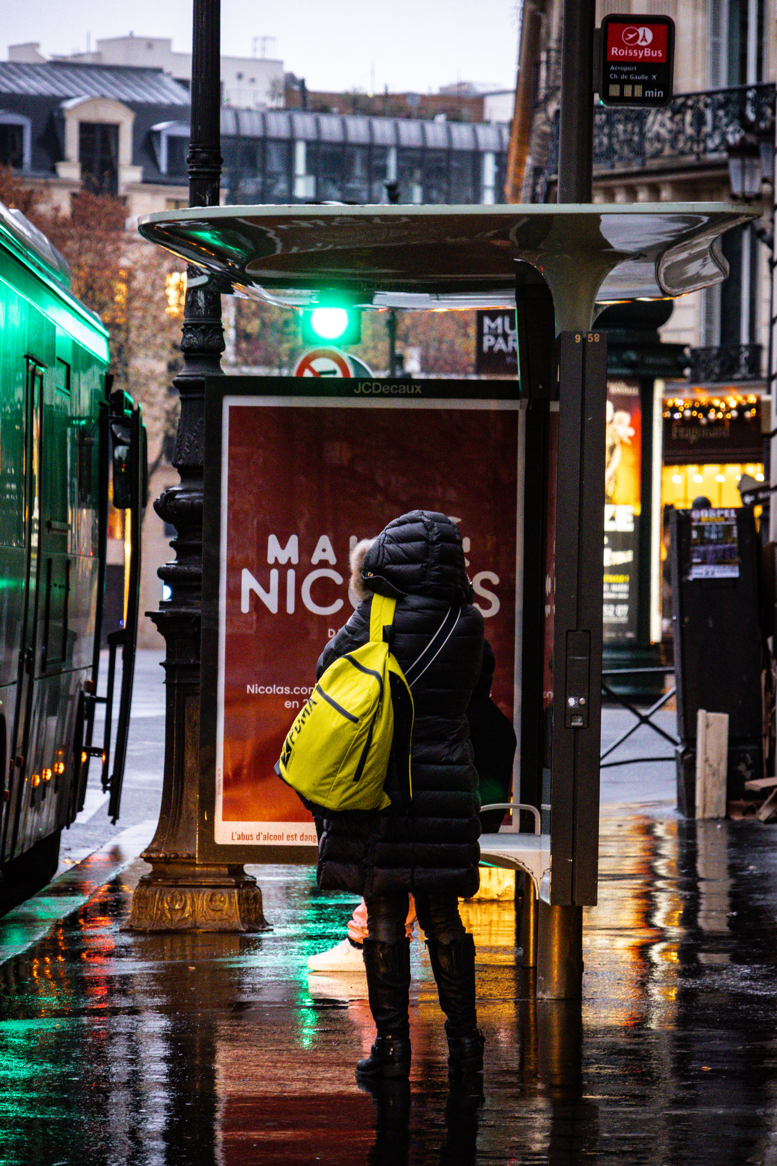 A person in a bright coat waiting beneath neon light on a rainy night.